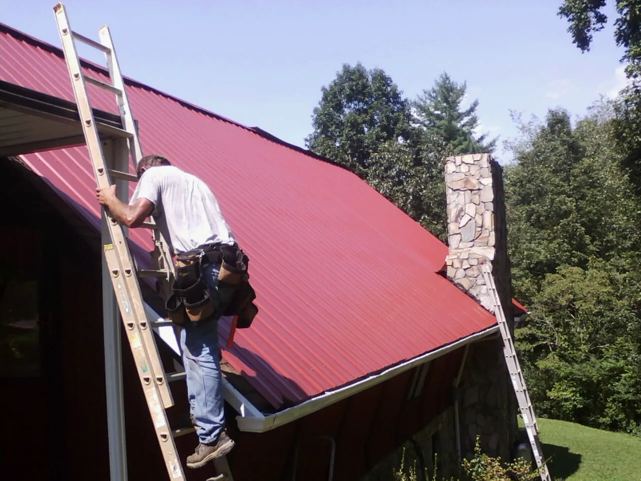 Roofer on a ladder beside a red metal roof and stone chimney — Soffit & Fascia Repair by Roofing Brothers in Gilberts